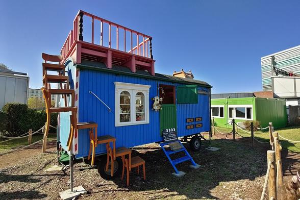 Ein bunt gestalteter, mobiler Bauwagen. Die Wände sind leuchtend blau, mit grüner Tür und weißen Fenstern. An der Seite führt eine Treppe aus übereinandergestapelten Holzstühlen nach oben zu einer Dachterrasse mit rotem Geländer. 