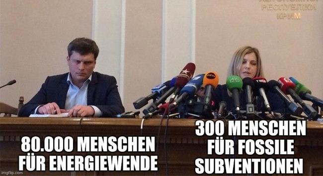 Zwei Personen sitzen bei einer Pressekonferenz hinter einem Tisch mit vielen Mikrofonen. Links sitzt ein Mann mit ernstem Blick, vor ihm steht der Text „80.000 Menschen für Energiewende“. Rechts sitzt eine Frau, die spricht, vor ihr steht der Text „300 Menschen für fossile Subventionen“. Das Bild stellt einen Vergleich zwischen unterschiedlich großer Unterstützung für zwei politische Themen dar.
