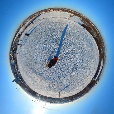 Ein kreisförmiges Bild im „Tiny Planet“-Stil zeigt eine schneebedeckte Landschaft unter einem klaren blauen Himmel. In der Mitte der Schneefläche befindet sich eine Person in dunkler Kleidung mit roten Akzenten. Ein langer Schatten der Person erstreckt sich über den unebenen Schnee nach oben rechts. Am Rand der kreisförmigen Landschaft sind Bäume, Gebäude und weitere Strukturen zu erkennen. Die Sonne ist am unteren linken Bildrand als heller Lichtpunkt sichtbar.
