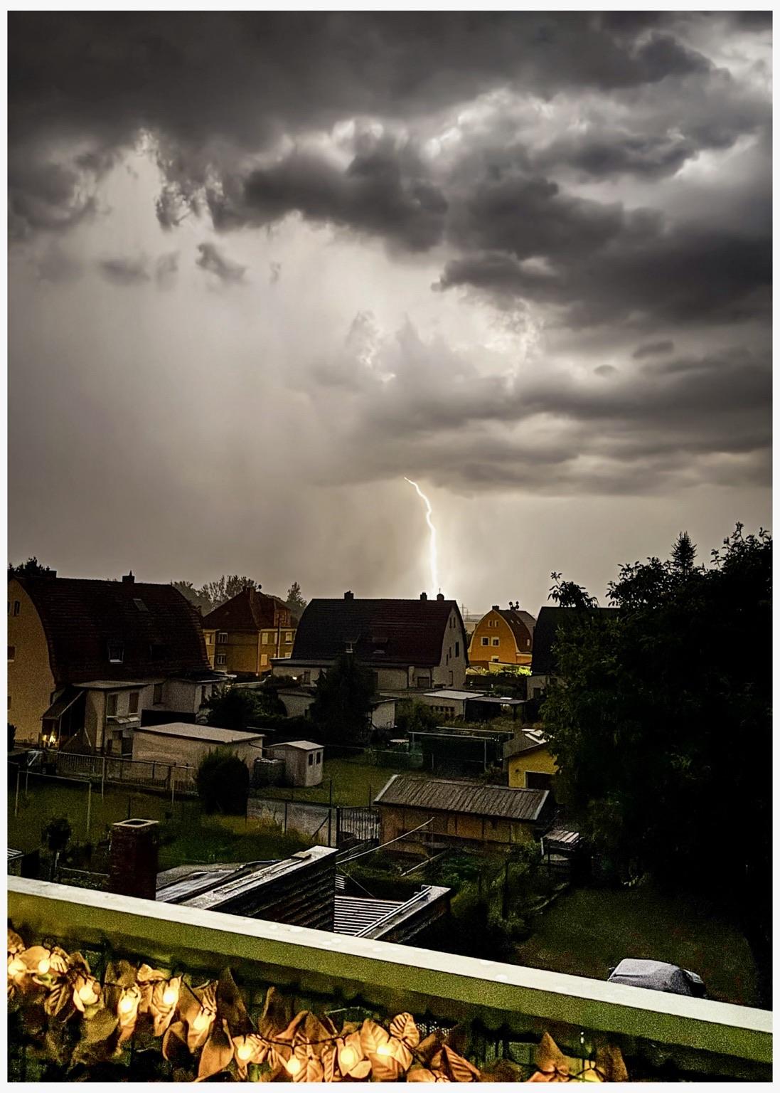 A dramatic storm scene with dark, cloudy skies and a bolt of lightning striking in the distance. In the foreground, residential houses and gardens are visible, with a balcony adorned with decorative lights.