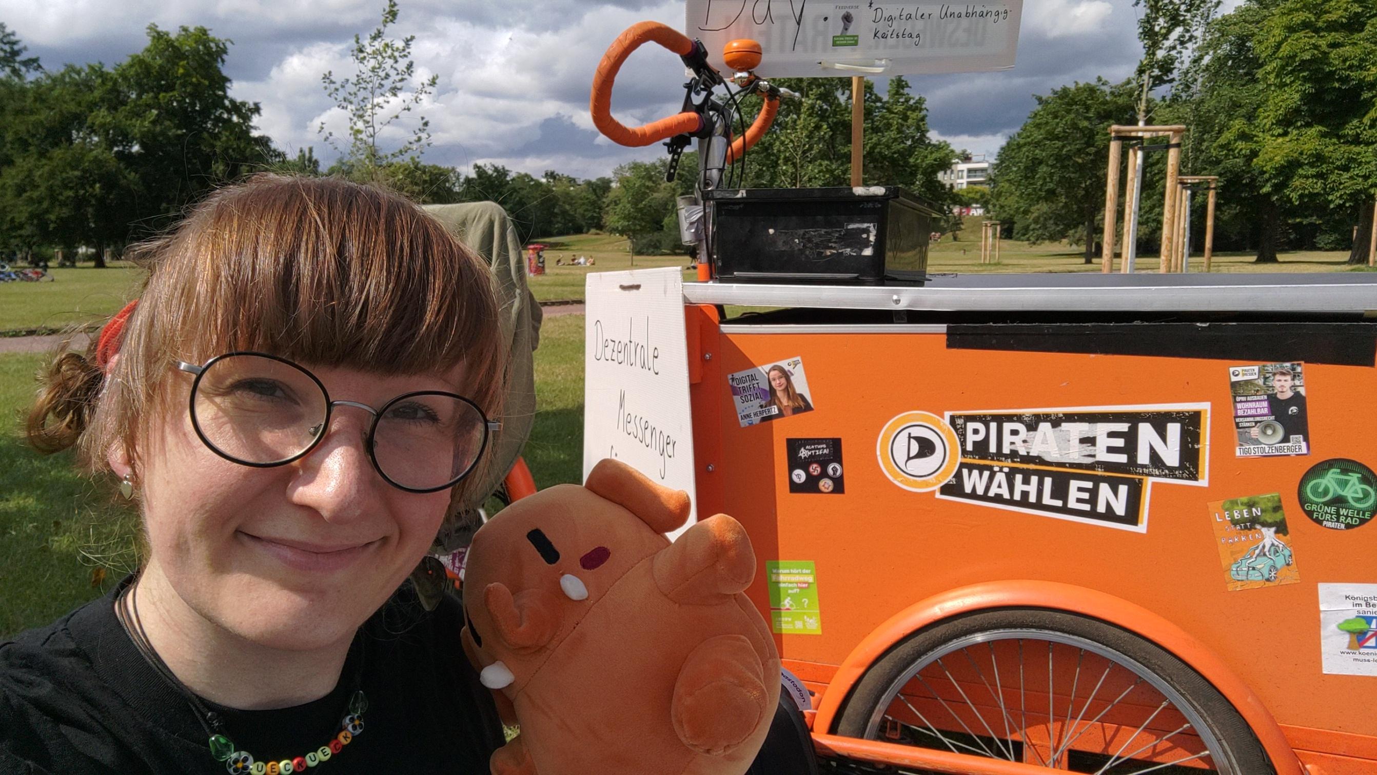 Ich, große runde Brille und Ponyfrisur im Park halte ein Madtodon-Plüschtier hoch und sitze neben einem Lastenrad.