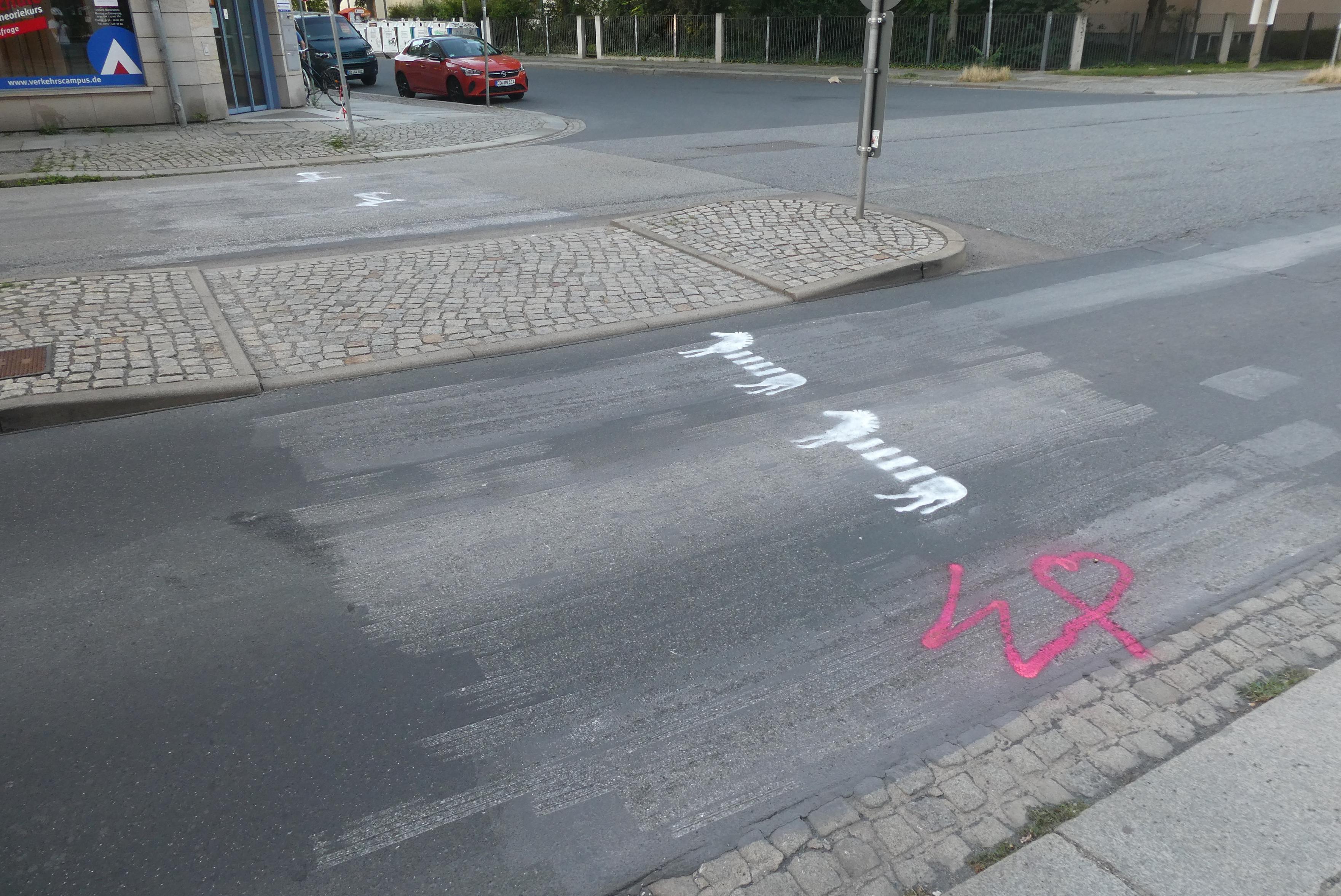 Die Pfotenhauerstraße mit Zebras bei Tag. An einem anderen Übergang mit abgesenkten Bordstein und Mittelinsel sind weiße Zebras auf die Fahrbahn gemalt, daneben in pink das Logo des Widerstandskollektivs: ein W, das in einem Herz endet. Der Straßenuntergrund ist grau und wie gebürstet, wo zuvor Zebrastreifen aufgemalt waren. Rechts im Bild ist der Kreuzungsbereich zur Arnoldstraße zu sehen.