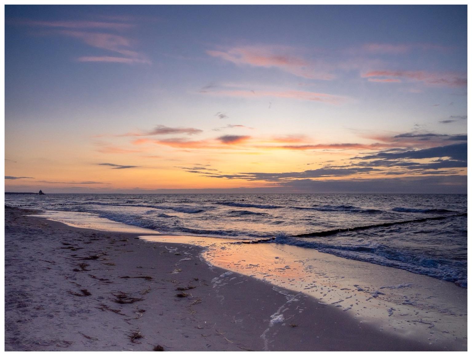 A serene beach scene at sunset, featuring gentle waves lapping against the shore. The sky is painted with hues of pink, orange, and purple, reflecting on the wet sand. A distant silhouette of a boardwalk appears on the horizon on the left.