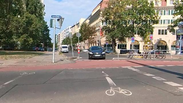 Straßenkreuzung in einer Stadt mit markiertem Radweg im Vordergrund. Links befindet sich eine Grünanlage mit hohen Bäumen, rechts stehen mehrstöckige historische Gebäude mit großen Fenstern. Mehrere Autos, ein Lieferwagen und abgestellte Fahrräder sind zu sehen. Ein Auto blockiert den Radweg. Die Straße ist sonnig, der Himmel teilweise bewölkt.