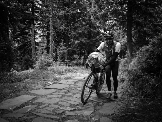 Black and white picture of a cyclist pushing his bike on a road with very large and uneven pavement. 