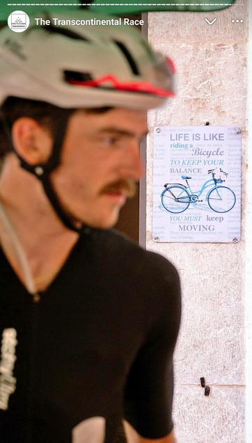 Unscharfer Radfahrer mit Helm im Vordergrund; im Hintergrund hängt ein Schild mit Fahrradmotiv und dem Spruch „Life is like riding a bicycle to keep your balance you must keep moving“ an einer hellen Wand.
