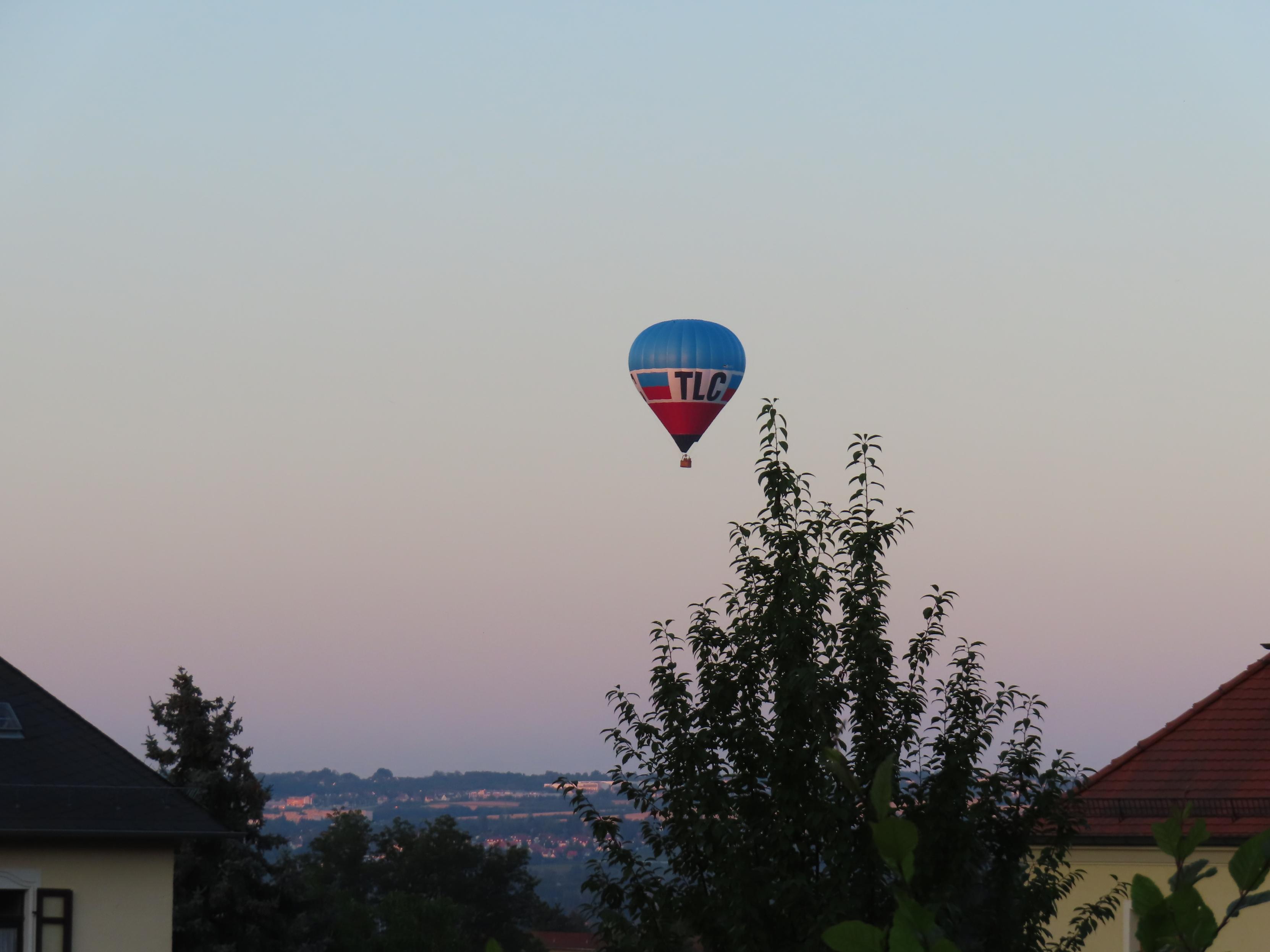 BlauWeißRoter Heißluftballon gegen 5.45Uhr am Himmel. 
In der Lücke zwischen zwei Dächern, über einem Baum