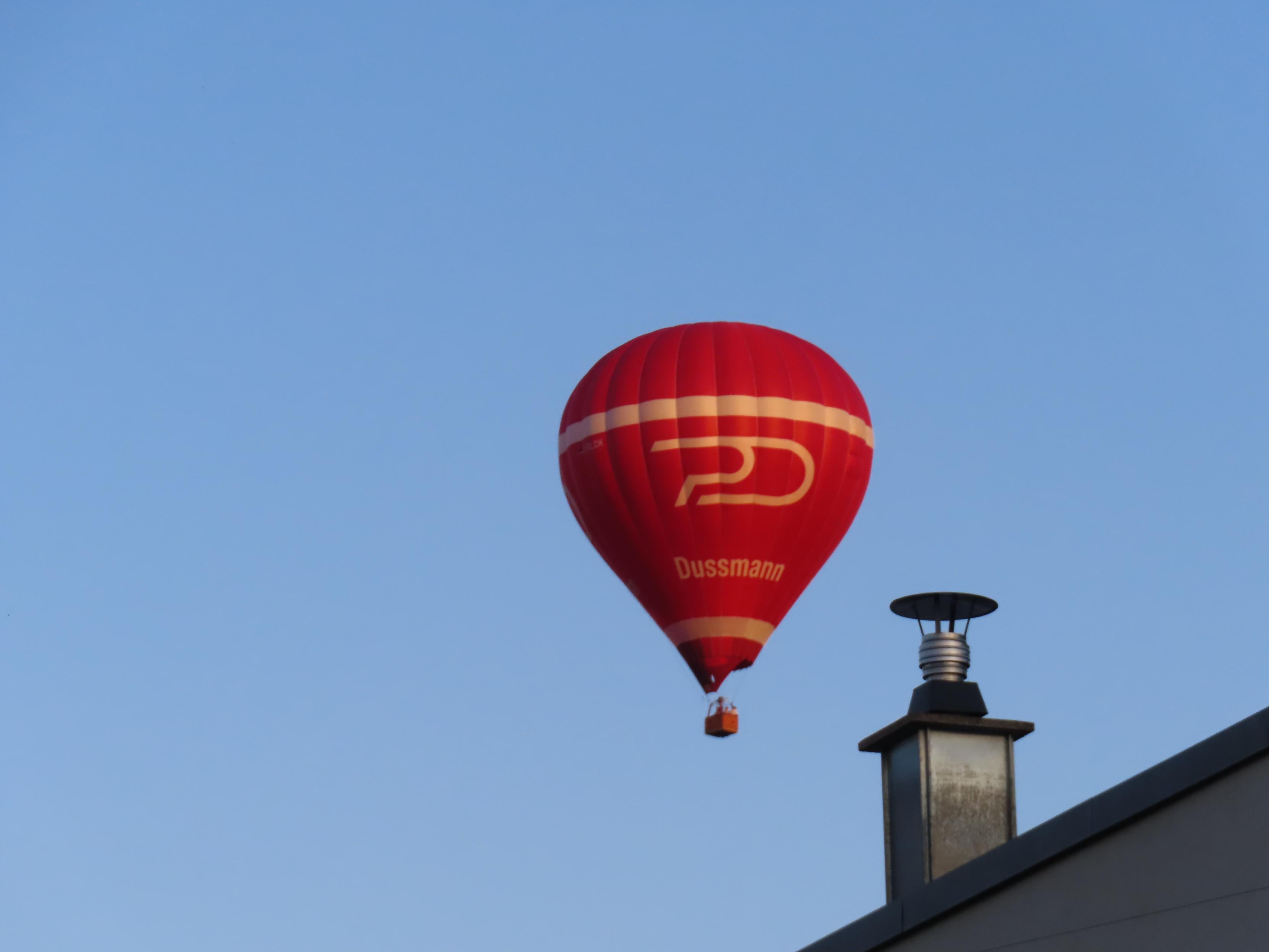 Roter Heißluftballon neben der Schornsteinanlage des Hauses gegenüber.
Blauer Himmel