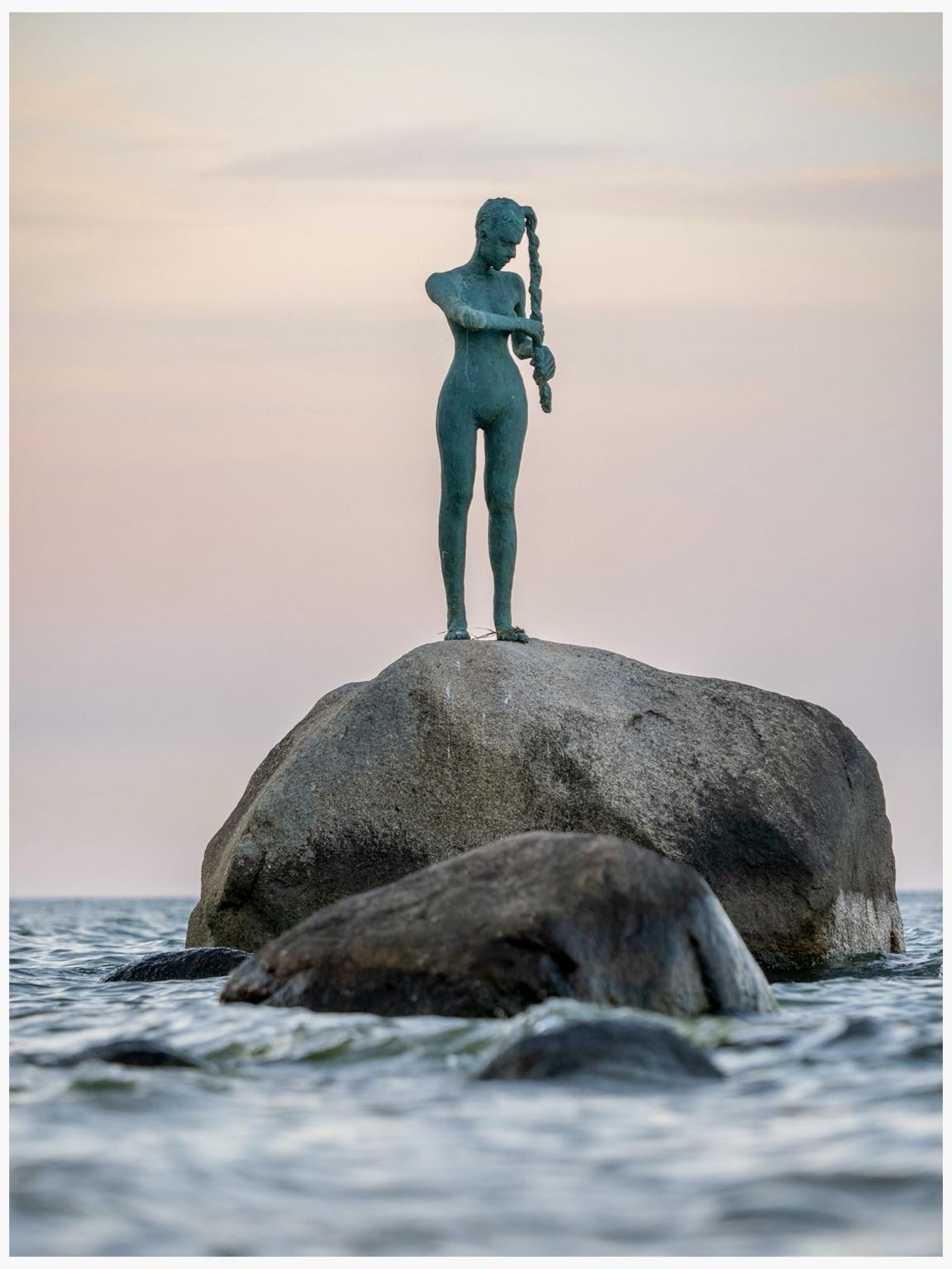 A bronze statue of a standing woman squeezing the water out  of her braid. It is perched on a large rock on the shore of the Baltic Sea. The background features a soft, pastel sky at dusk.