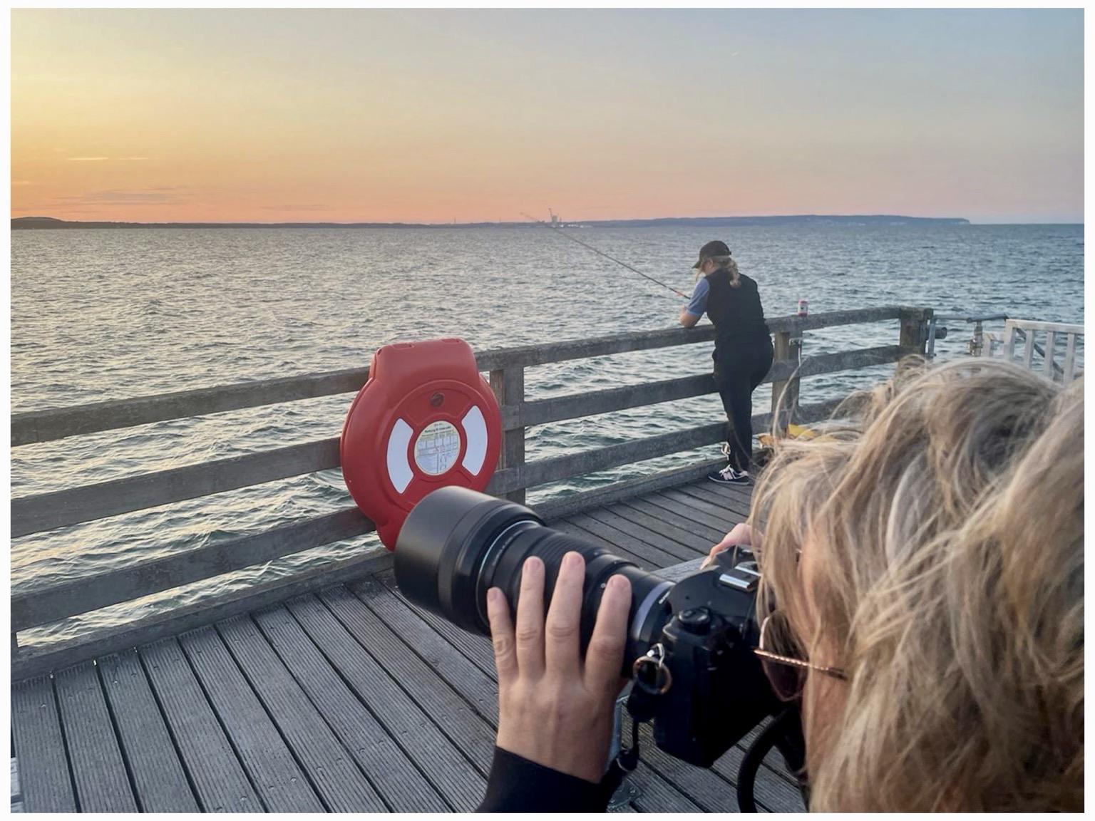 A woman with a camera focuses on the scene while another woman fishes off a wooden pier at sunset. A red life ring is mounted nearby. The water reflects the warm colors of the sky.