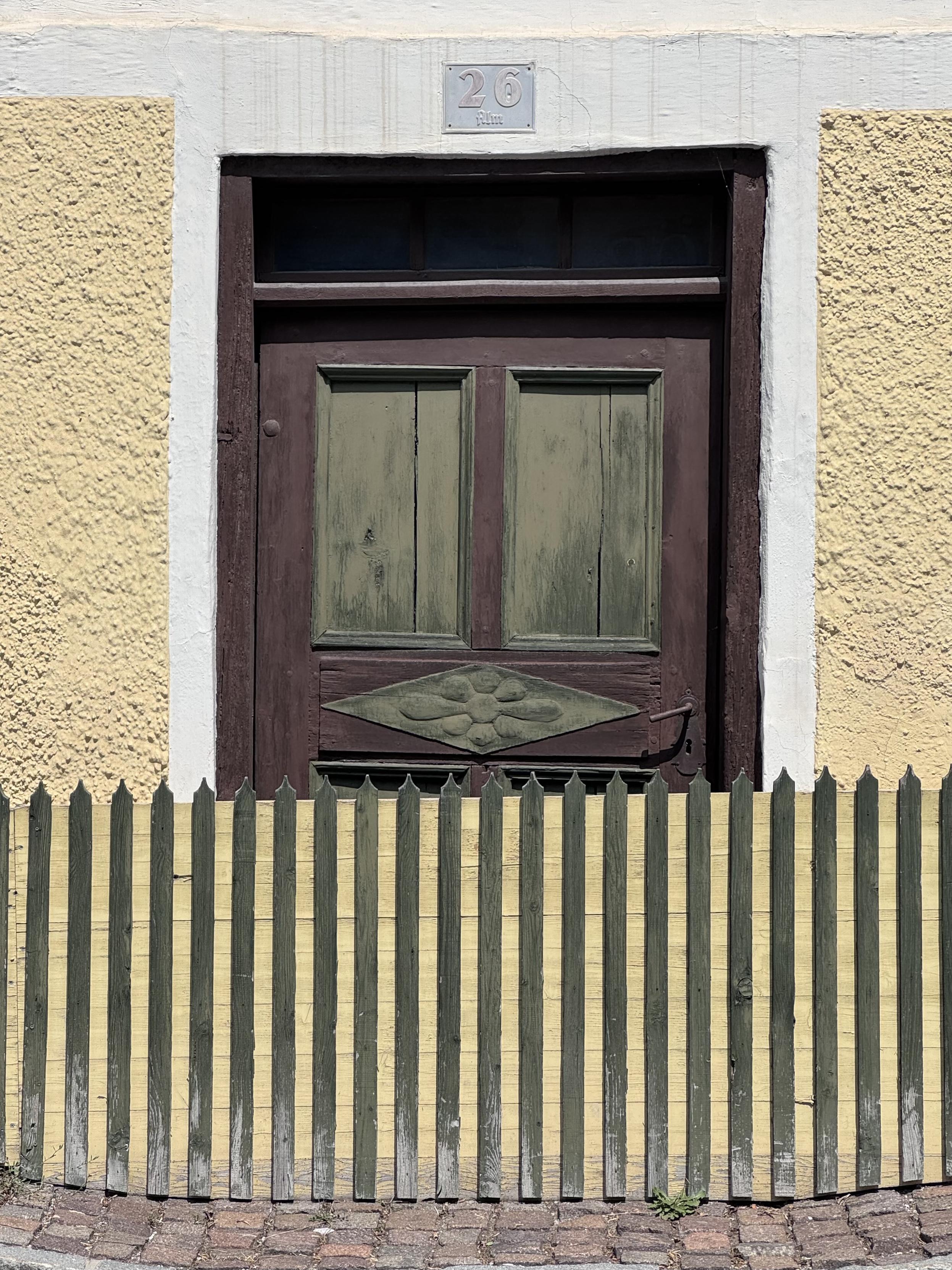 Eine alte Eingangstür in einer gelben Fassade. Mit der Mauer direkt abschließend eine ArtZaun, der ein Drittel der Tür verdeckt und die Benutzung der Tür unmöglich macht