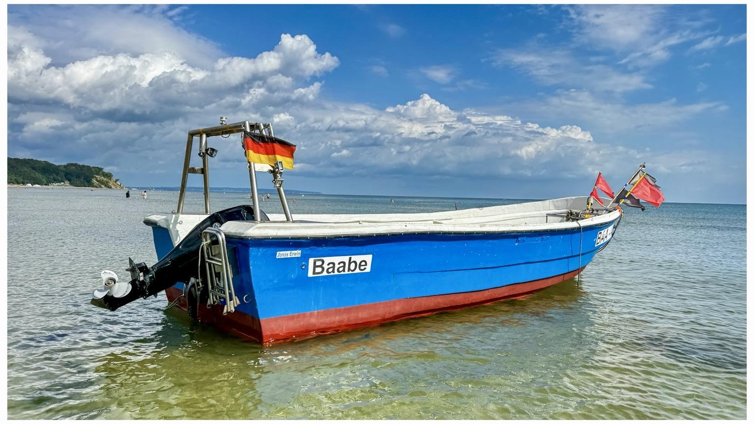 A blue fishing boat named "Baabe" is anchored in shallow water in the Baltic Sea, with a German flag and red flags waving above. The background features a clear sky with scattered clouds and a shoreline in the distance.