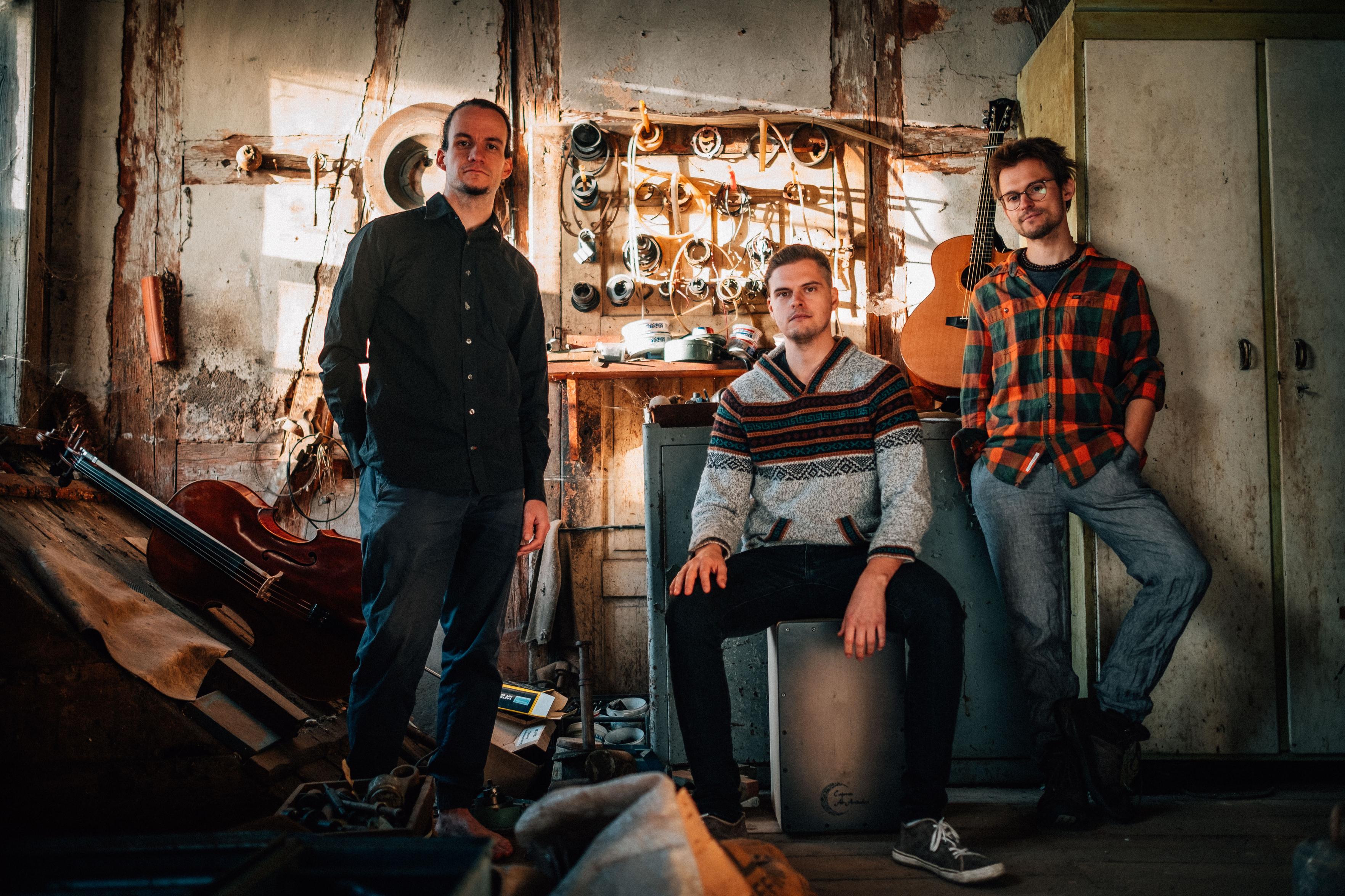 Die drei Musiker der Band stehen in einer Werkstatt. Im Bild zusätzlich ein Cello und eine Gitarre.
