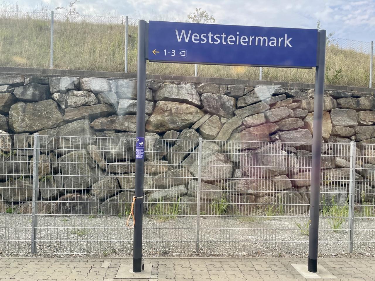 Blick aus dem Zugfenster auf eine blaue Tafel, welche mit Weststeiermark beschriftet ist, dahinter eine Steinmauer und Wiese