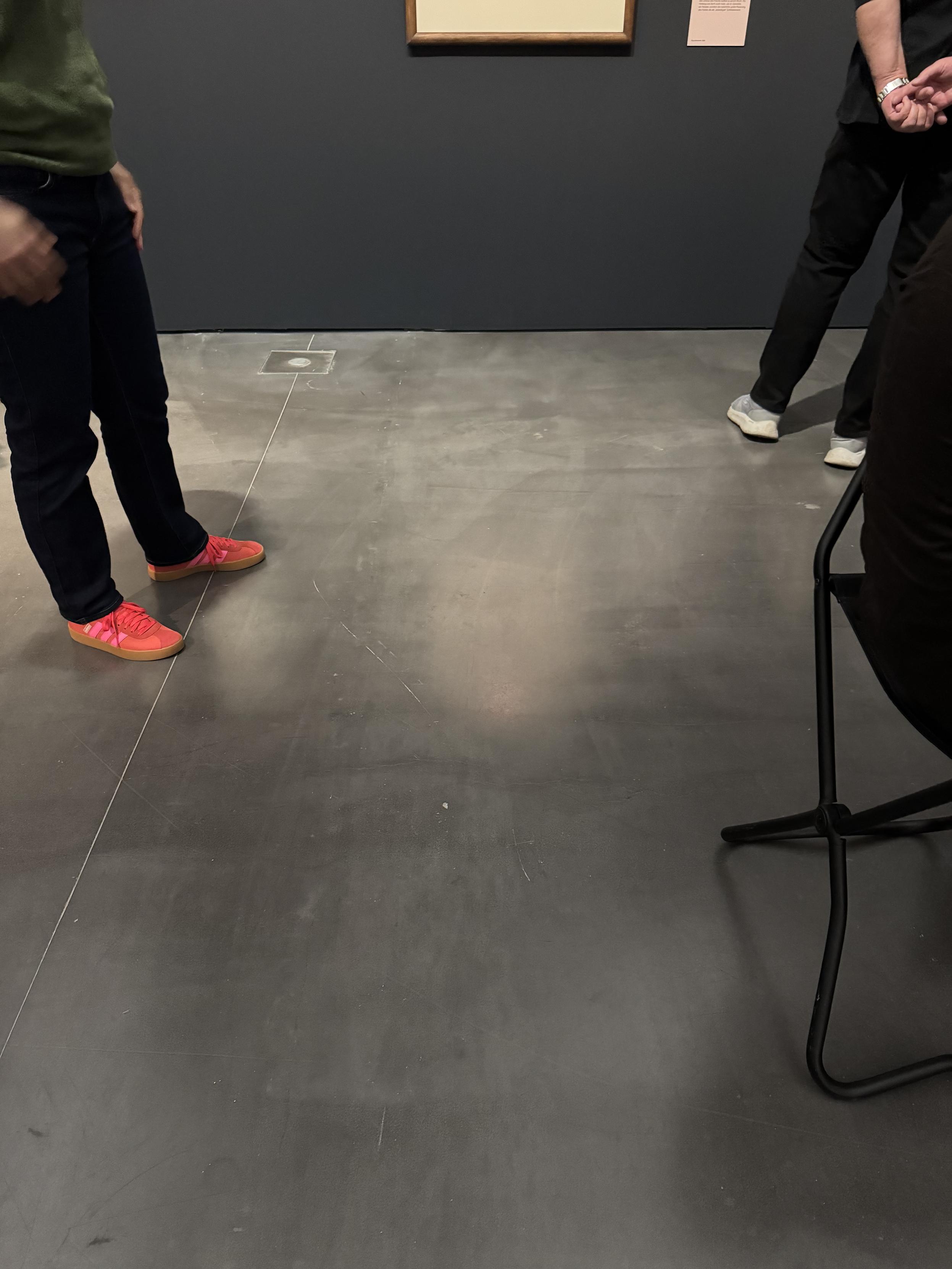 Blick auf einen dunkelgrauen Fußboden und ein Stück dunkelgraue Wand. Links Hosenbeine und rote Sportschuhe einer Person, rechts im Bild ein Hocker/Klappschemel und die Hosenbeine und weiße Schuhe einer weiteren Person. Am oberen Bildrand angeschnitten zwei Bilderrahmen.