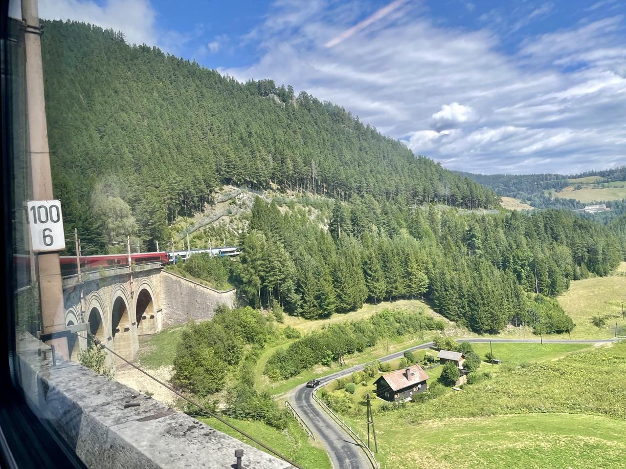 Blick aus dem Zugfenster:
unser Railjet fährt gerade über ein Viadukt, ein weiß-blauer Zug begegnet uns und es gibt Aussicht auf eine bewaldete, bergige Landschaft und grüne Wiese, eine Straße, ein paar Häuser und alles bei blauem Himmel, Sonnenschein und weißen Wolken