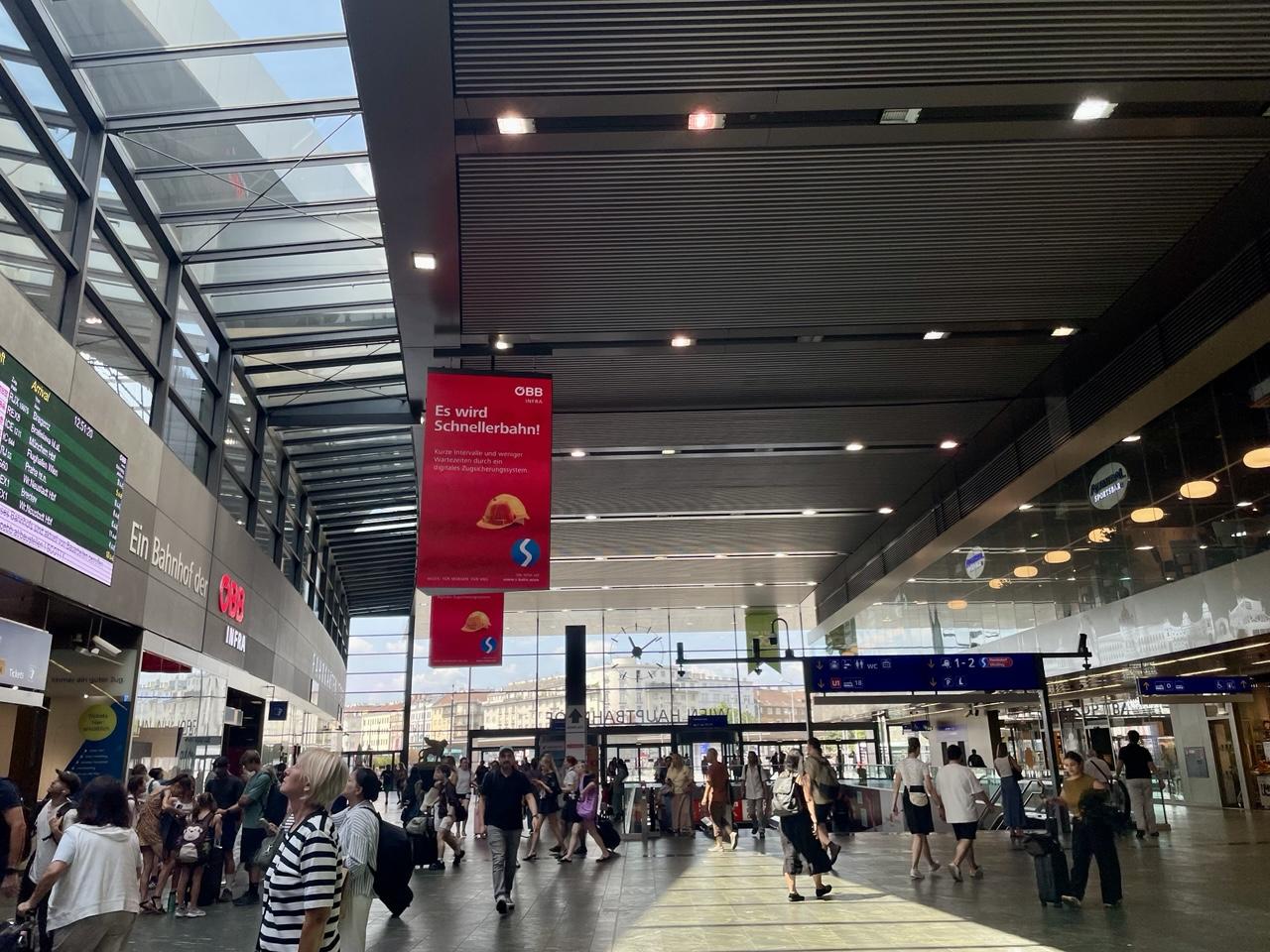 Große Halle mit Glasfront, allerhand Leute, von der Decke rote Plakate der ÖBB Infra:

Es wird
Schnellerbahn!

Kurze Intervalle und weniger Wartezeiten durch ein digitales Zugsicherungssystem

Ein gelber Bauhelm ein weißes S auf blauem Kreis.

Heute
Für Morgen
Für uns

Alle Infos auf
www.s-bahn.wien