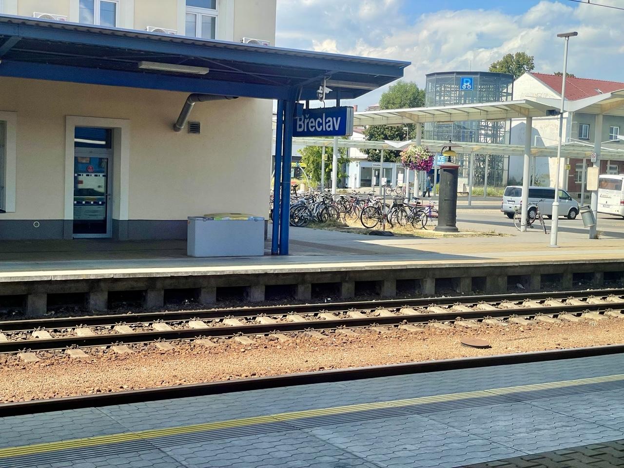 Blick aus dem Zugfenster: Gleise, die Ecke eines Bahnhofsgebäudes, an der das Schild Břeclav hängt, ein mit Blumen geschmückter Fahrradparkplatz, im Hintergrund ein Fahrradparkhaus