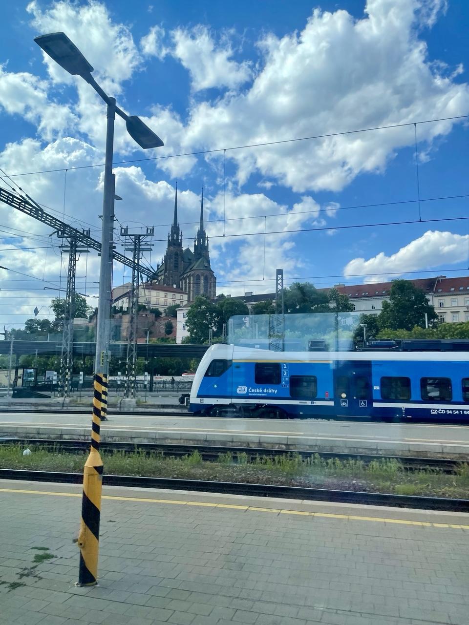 Blick aus dem Zugfenster: Bahnsteige, Gleise, eine Laterne ein blau weißer Zug dahinter Häuser und die Kathedrale St. Peter und Paul unter blauem Himmel mit weißen Wolken