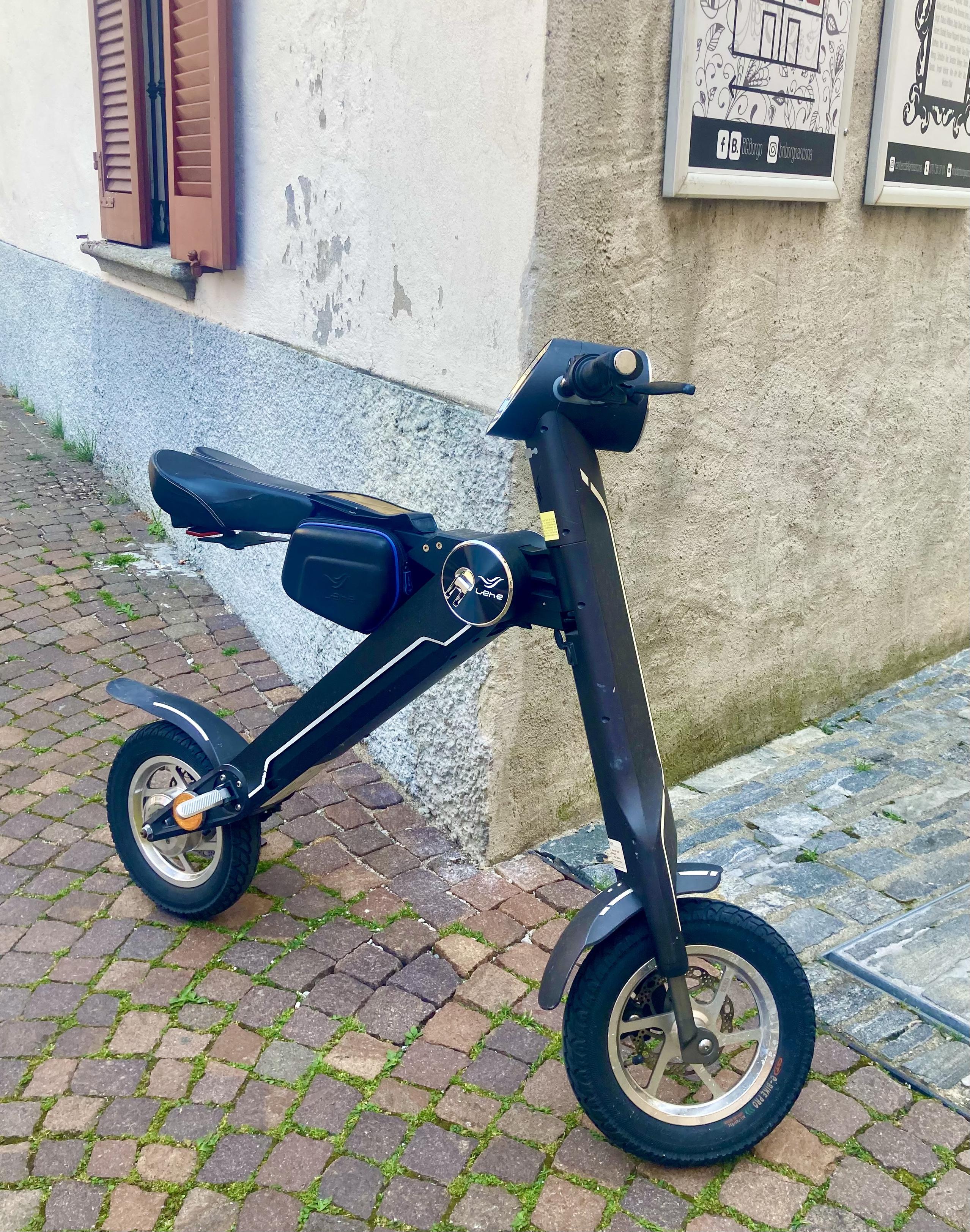 Ein  sehr modern aussehendes schwarzes elektrisches Laufrad, an eine Hausecke gelehnt