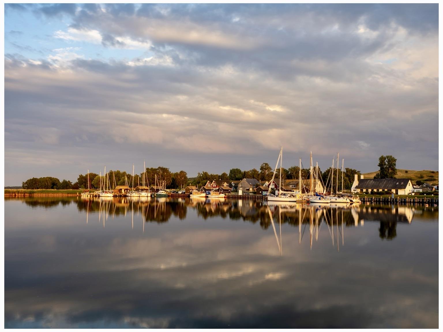 A serene waterfront scene featuring a collection of boats docked at a small harbor. The calm water reflects the colorful houses and sailboats, surrounded by trees and under a partly cloudy sky. The image captures a peaceful, picturesque coastal atmosphere.