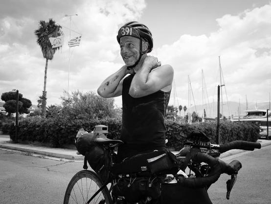 Schwarz-weiß-Foto eines lächelnden Radfahrers mit Helm und Rennrad, der an einer Uferpromenade steht. Im Hintergrund sind Palmen, Boote und Segelmasten vor einem wolkigen Himmel zu sehen.

