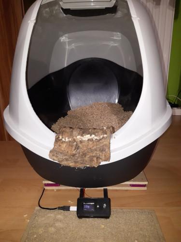 A white litter box with a piece or cork bark on the entrance. Under the litter box there are two wooden boards with load cells inbetween and a black 3d printed case with a small OLED display and a control knob