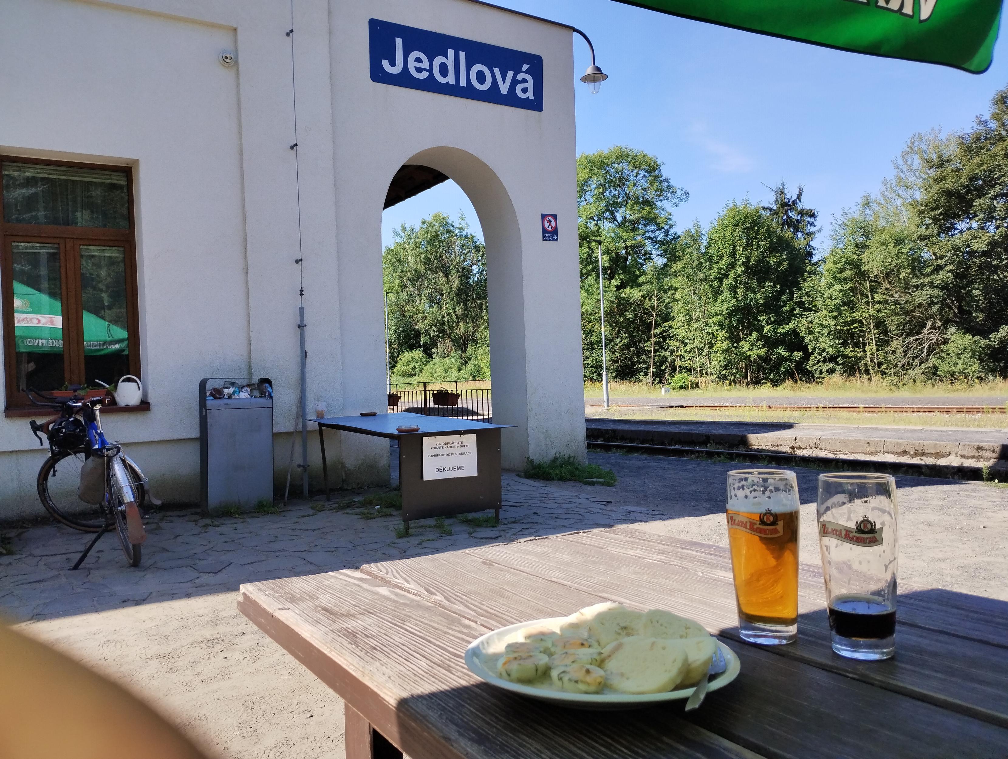 Bahnhofsgebäude mit seitlichem Torbogen und Aufschrift "Jedlowa", davor ein Fahrrad, Im Vordergrund ein Teller mit Eiern und Knödeln, eine Kofola und ein Bier.