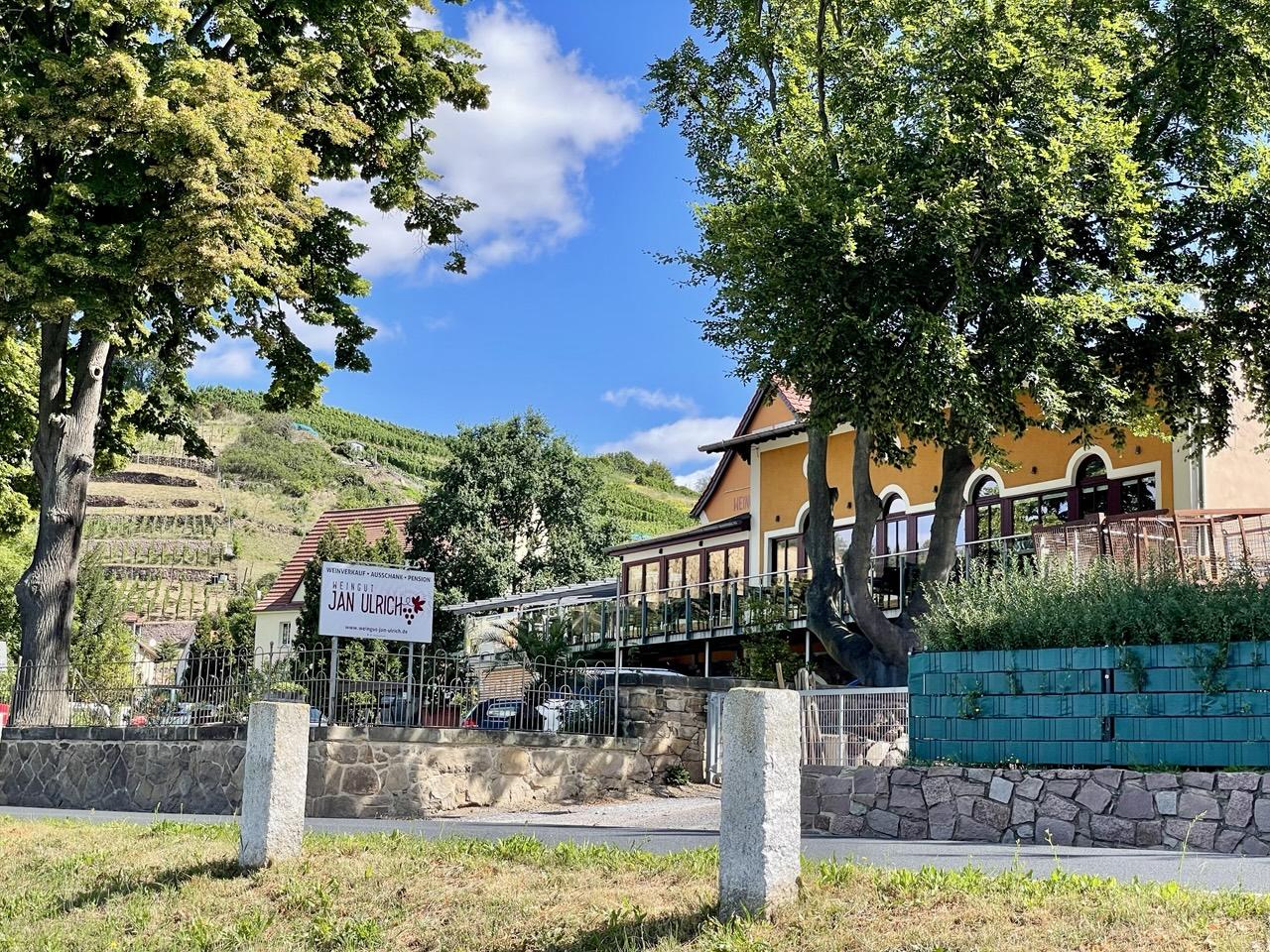 Zwei Steinpfosten an der Straße, auf der anderen Straßenseite ein Schild:
Weinverkauf Ausschank Pension
Weingut
JAN ULRICHS