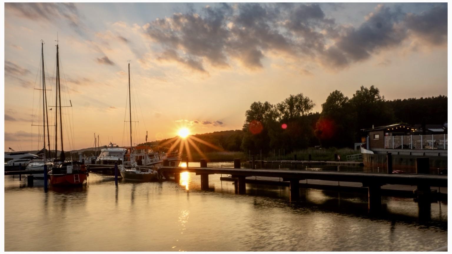 A scenic sunset over a marina, featuring several boats docked alongside a wooden pier. The sky is filled with colorful clouds, reflecting the golden hues of the setting sun on the calm water. In the background, trees line the shore, and a building