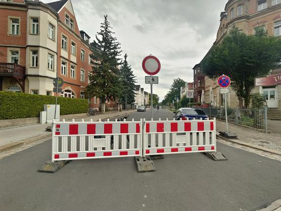 Eine innerstädtische Straße mit historischen Wohn- und Geschäftsgebäuden ist durch rot-weiße Absperrgitter für den Verkehr gesperrt. Dahinter stehen Verkehrsschilder, Fahrräder und ein geparktes Auto; im Hintergrund führt die Straße zwischen Bäumen und Gebäuden weiter.