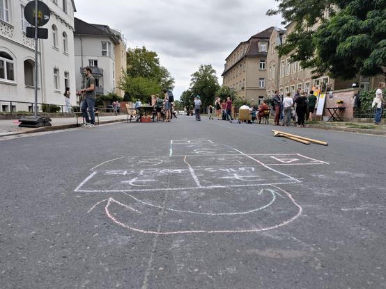 Auf einer gesperrten Wohnstraße spielen und treffen sich mehrere Menschen. Im Vordergrund ist mit bunter Kreide ein großes Hüpfspiel auf die Fahrbahn gemalt; im Hintergrund sitzen und stehen Menschen zwischen Wohnhäusern, Tischen und Stühlen.
