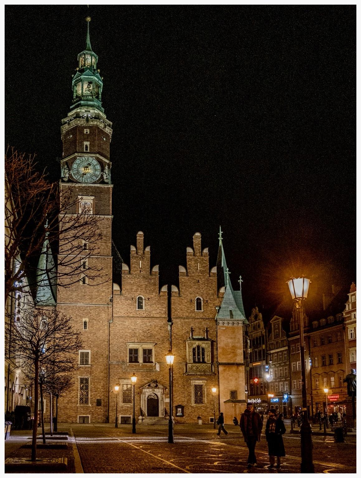 A historic building with a tall clock tower, illuminated at night, surrounded by cobblestone streets and street lamps. People are walking nearby.