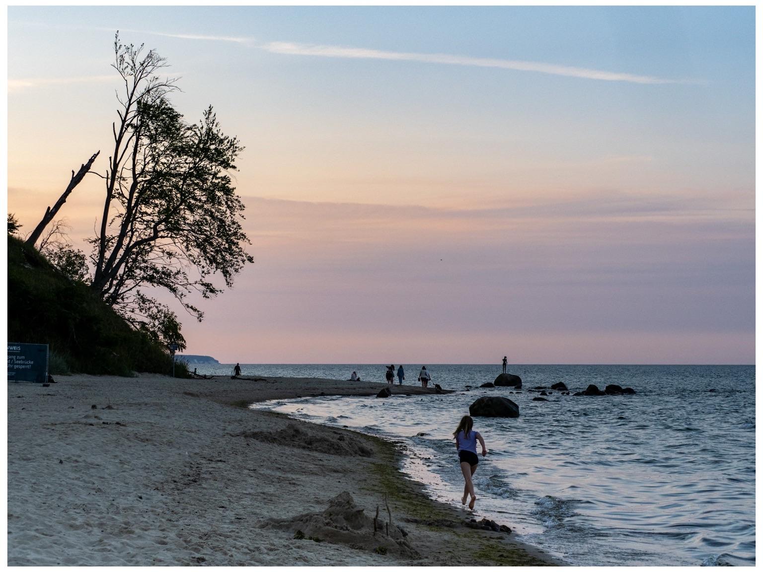 A beach scene at sunset. A girl running along the beach. In the background, people are walking on the shore and standing on rocks. Silhouettes of trees on the left and a vibrant pastel can be seen in the background.