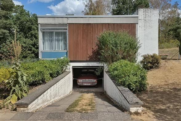 Ein Gebäude mit einer Einfahrt, die zu einer Garage unter dem Straßenniveau Straßenniveau führt. Das obere Stockwerk zeigt links ein Fenster mit weißen weißen Vorhängen und einer blauen Fläche darunter, während die rechte Seite mit vertikalen braunen Holzlatten bedeckt ist. Ein weißer Ziegelwandabschnitt bildet den rechten Abschluss des Gebäudes. Eine gepflasterte Rampe führt zwischen grünen Sträuchern hinunter zu einem dunklen Auto in der Garage.