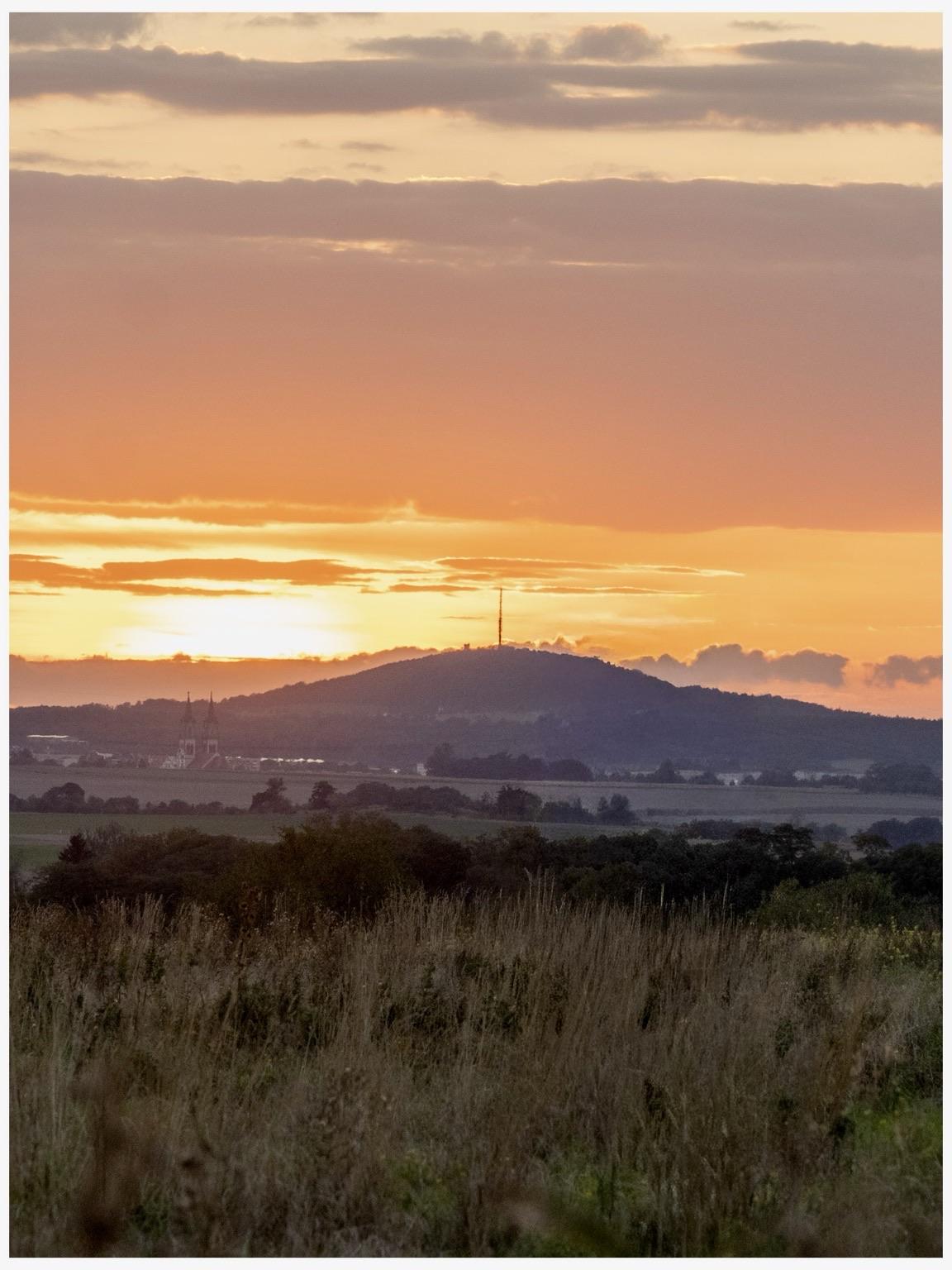 Bild hochkant: In der Ferne ein Hügel mit Sendemast. Dahinter orangefarbener Sonnenuntergang mit ein paar dunklen Wolken. Davor Felder und Gebüsch.