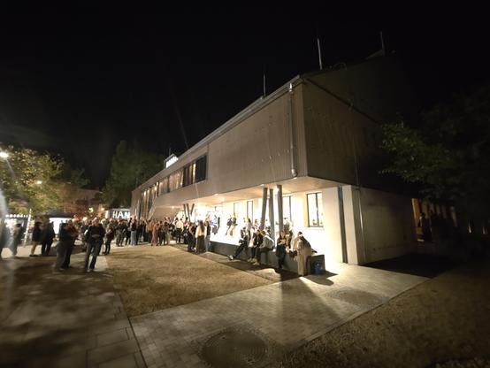 Ein Nachtfoto zeigt ein zweistöckiges Gebäude mit einer hellen, vertikal strukturierten Fassade und einer Menschenmenge. Unter einem beleuchteten Vordach, das von Säulen gestützt wird, sowie auf einer Fläche mit Schotter stehen viele Personen. Im Vordergrund befindet sich ein gepflasterter Weg. Auf der linken und rechten Seite des Gebäudes sind dunkle Bäume zu sehen. Die Szene wird durch künstliche Lichtquellen beleuchtet, während der Himmel schwarz ist.