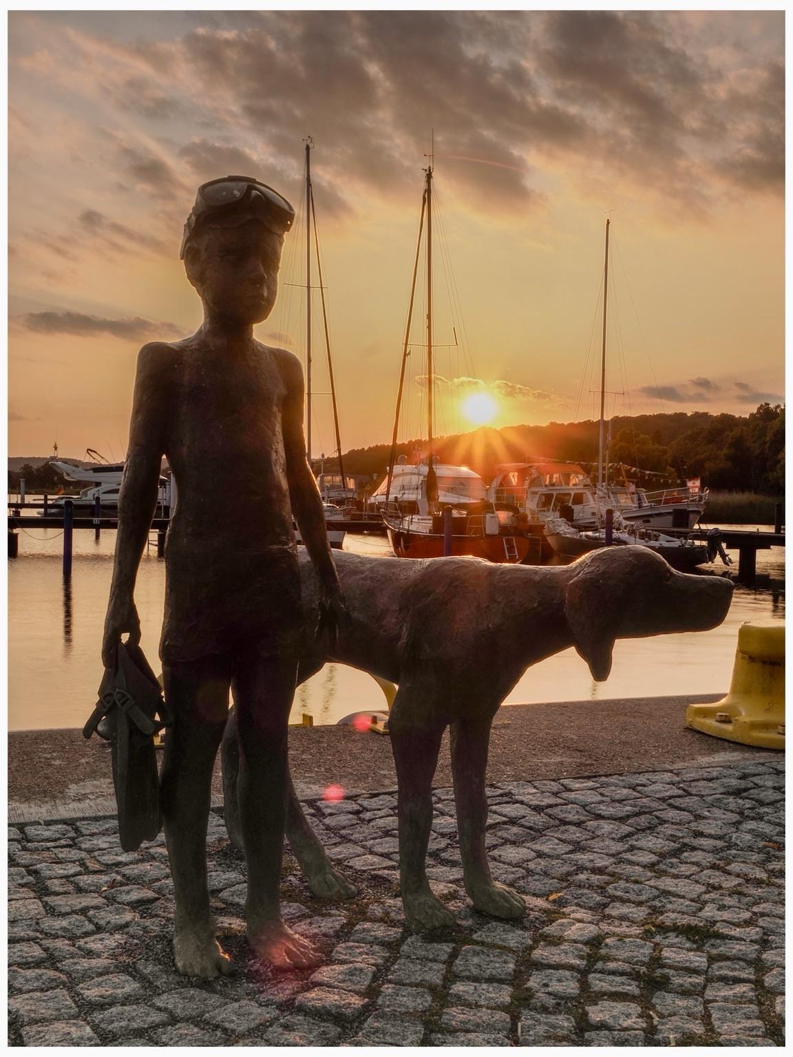 Eine Skulptur im Vordergrund zeigt einen Jungen mit hochgeschobener Taucherbrille und Flissen in der Hand. Links neben ihm ein labradorähnlicher Hund. 
Beide stehen an der Wasserkante zu einem Hafen für Segelboote. Im Hintergrund geht hinter einem waldigen Hügel die Sonne unter. Die Szene ist in goldgelbes Abendlicht getaucht.